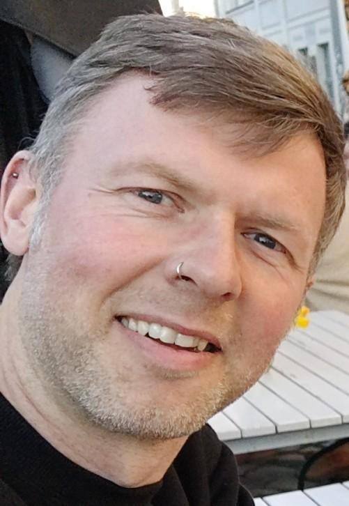 Christian schaut seitlich in die Kamera. Er hat kurzes grau-braunes Haar und einen Dreitagebart. Er trägt einen Nasenring und einen kleinen Stecker oben im rechten Ohr.