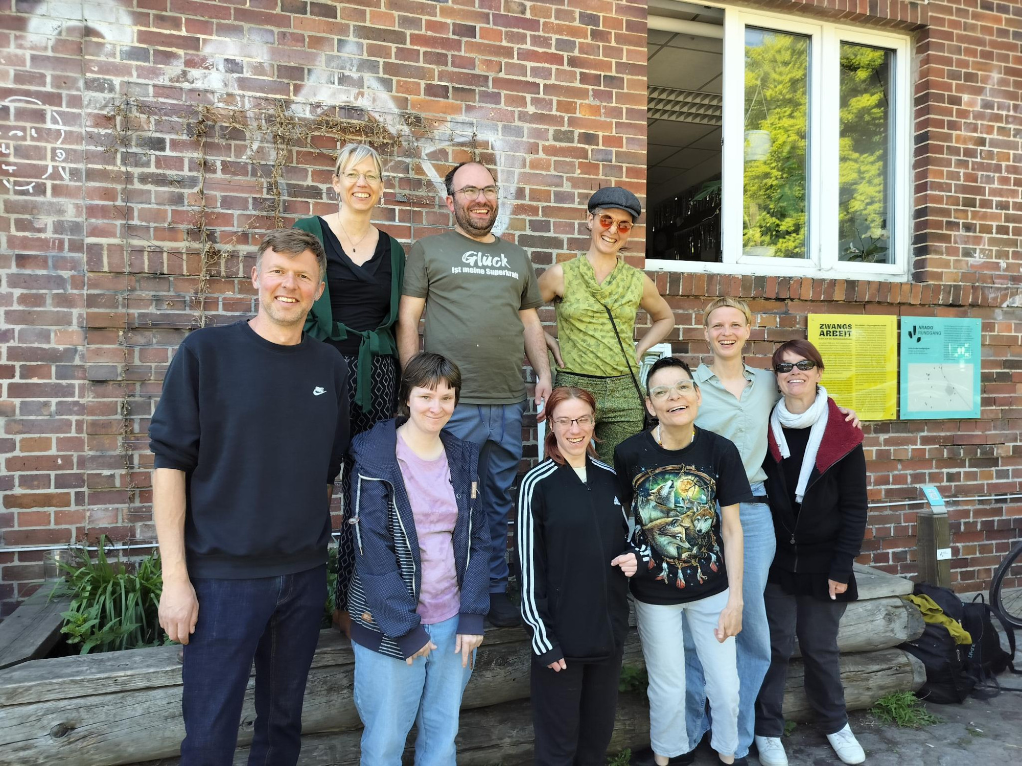 Gruppenfoto Arado Weg: von links nach rechts: Christian Raschke, Maxie Karius, Janiene Gaebel, Richard Kunkel, Cindy Knoke, Jolika Sudermann-van den Berg, Christine Schuster, Manuele Schulz, Anke Landeck. Alle lachen und stehen vor einer roten Hauswand aus Backsteinen.