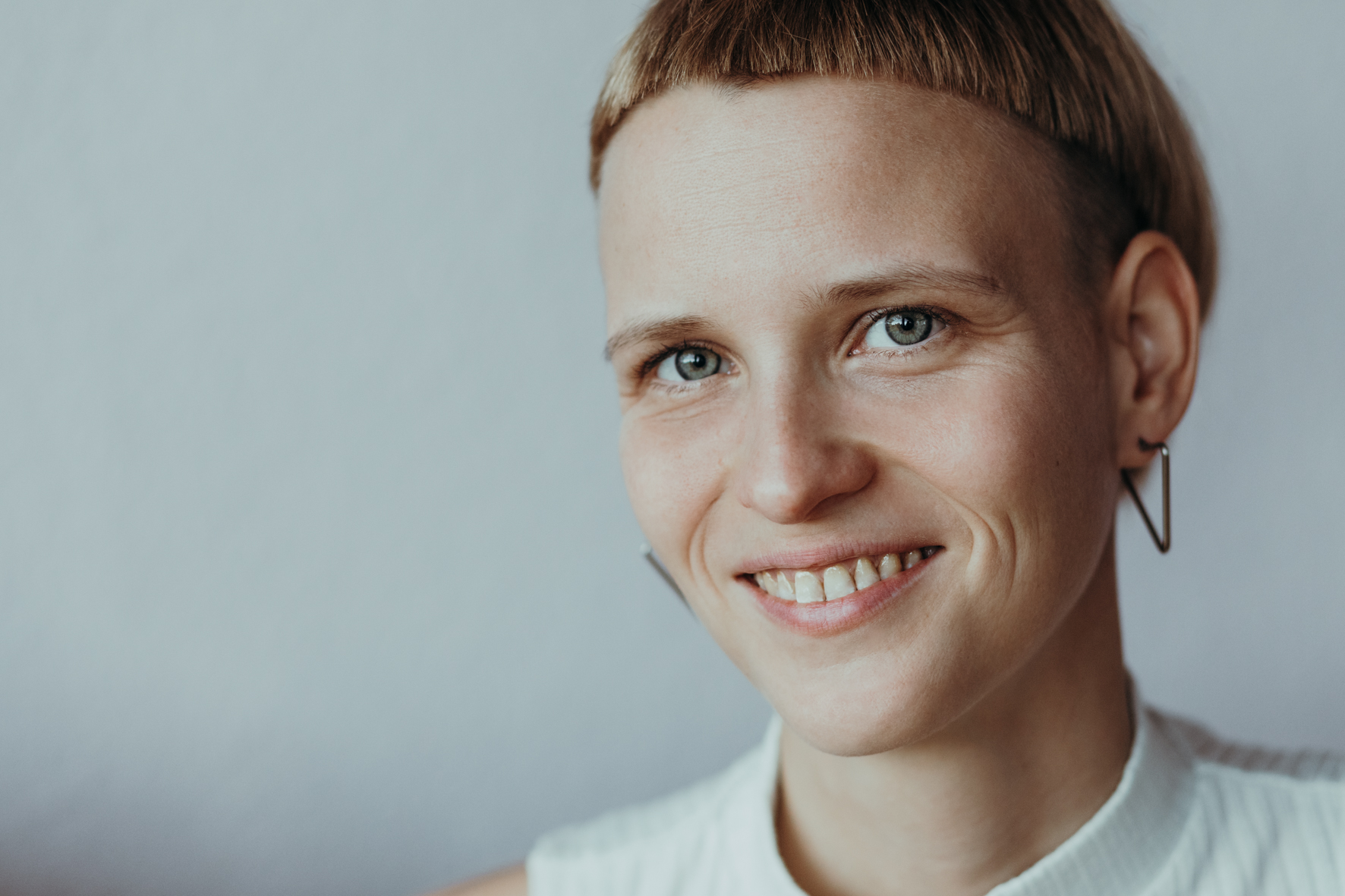 Manu hat kurze blonde Haare und einen sehr geraden Pony. Sie trägt ein weißes Oberteil und dreieckige Ohrringe. Sie guckt schräg in die Kamera und lächelt.